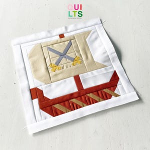 May include: A square fabric quilt block featuring a pieced design of a Viking ship with a white sail, a tan mast, and a red hull with gold diagonal stripes. The block is framed by a white border and includes the text QUILTS AND NICE STUFF at the top.
