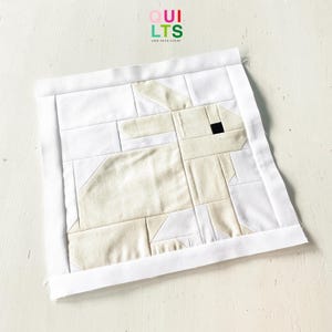 May include: A handmade fabric bunny quilt block. The bunny is made of beige and white fabric pieces, with a black square for the eye. The block is framed with a white border. The text "QUILTS AND NICE STUFF" is in the upper right corner.