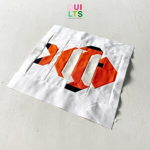 Könnte beinhalten: Ein weißer Stoffquadrat mit einem bunten Fischdesign aus Stoffresten. Der Fisch ist orange, weiß und schwarz. Der Stoffquadrat hat den Text "QUI LTS AND NICE STUFF" in der oberen linken Ecke.
