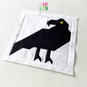 Könnte beinhalten: Ein handgefertigter Quiltblock mit einem geometrischen Design eines schwarzen Raben auf weißem Hintergrund. Der Rabe hat ein gelbes Auge. Der Block ist mit einem weißen Rand eingefasst. Der Text "QUILTS" und "AND NICE STUFF" befindet sich in der oberen linken Ecke.