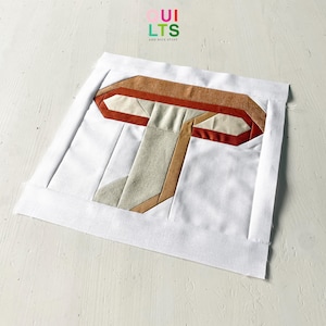 Könnte beinhalten: Ein Stoff-Pilz-Quiltblock mit weißem Hintergrund und einem Pilzdesign in Braun-, Beige- und Cremetönen. Der Block ist von einem weißen Rand umgeben. Der Text "Quilts and Nice Stuff" befindet sich in der oberen rechten Ecke.