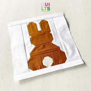 May include: A handmade quilt block with a brown bunny design and a white tail, set against a white background. The bunny is made of geometric shapes. The text "QUILTS AND NICE STUFF" is in the top right corner.