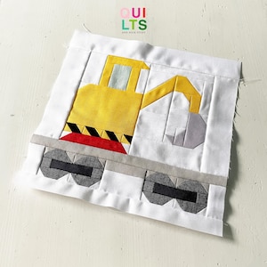 Könnte beinhalten: Ein handgefertigter Quiltblock mit einem gelben Baufahrzeug mit einem schwarz-roten Design. Der Block ist vor einem weißen Hintergrund platziert, mit dem Text "QUILTS AND NICE STUFF" in der oberen linken Ecke.