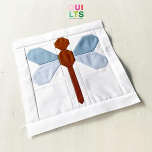 May include: A white fabric square featuring a dragonfly design. The dragonfly has brown body segments, light blue wings, and a brown tail. The words "QUILTS" and "AND NICE STUFF" are in the upper right corner.
