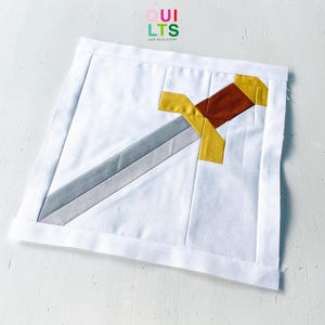 May include: A square fabric quilt block featuring a pieced sword design in shades of grey, brown, and yellow against a white background. The sword is positioned diagonally across the center. The text QUILTS AND NICE STUFF is printed at the top.
