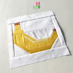 Könnte beinhalten: Ein Stoffblock mit einem geometrischen Bananen-Design in Gelb und Braun, vor einem weißen Hintergrund und von einem weißen Rand umgeben. Der Text "Quilts and Nice Stuff" ist in der oberen rechten Ecke sichtbar. Das Design erinnert an einen Quilt.