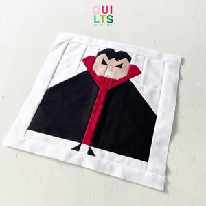Könnte beinhalten: Ein Stoffblock mit einem Vampir-Design. Der Vampir hat einen schwarzen Umhang, einen roten Kragen und ein blasses Gesicht mit ernstem Ausdruck. Der Block ist von weißem Stoff umrandet und enthält den Text "QUILTS AND NICE STUFF".