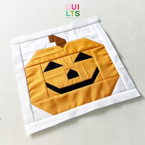 Könnte beinhalten: Quadratische, gesteppte Halloween-Dekoration mit einem lächelnden Kürbisgesicht. Der Kürbis besteht aus orangefarbenem Stoff mit schwarzen Augen, Nase und Mund. Der Hintergrund ist weiß mit einem weißen Rand. Der Text "QUILTS AND NICE STUFF" befindet sich oben rechts.