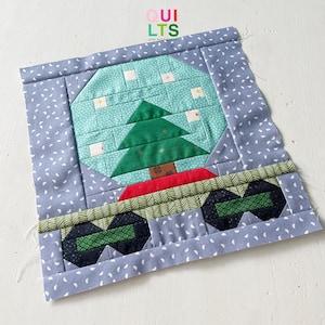May include: A blue and white fabric block with a green Christmas tree inside a blue and white snow globe. The snow globe is sitting on a green and red base with black and green wheels.