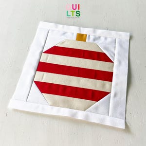 Könnte beinhalten: Ein quadratisches, gestepptes Weihnachtsornament-Design. Das Ornament hat ein rot-cremefarbenes Streifenmuster mit einem goldenen Oberteil, das von einem weißen Rand umrahmt wird. Der Text "Quilts and Nice Stuff" befindet sich in der oberen rechten Ecke.