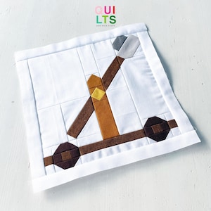 Op de afbeelding: Een vierkant quiltblok van stof met een samengesteld ontwerp van een katapult in bruine, beige en grijze tinten op een witte achtergrond. De katapult heeft een centrale verticale paal, een diagonale arm en twee donkerbruine wielen. Tekst: QUILTS AND NICE STUFF.