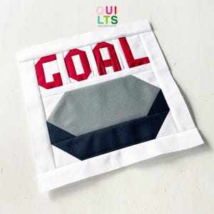 May include: A handmade quilt block with the word "GOAL" in red letters. Below the word is a gray and black geometric design. The block is framed with a white border. The text "QUILTS AND NICE STUFF" is in the upper right corner.