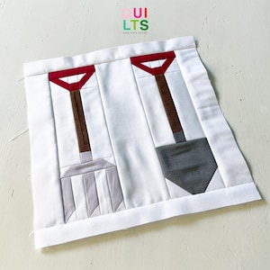 Peut inclure: Un bloc de courtepointe en tissu carré avec un fond blanc et deux outils de jardinage appliqués au centre. Un outil est une fourche gris clair et l'autre est une pelle gris foncé, tous deux avec des manches marron et des poignées rouges en Y. Le texte indique QUILTS.
