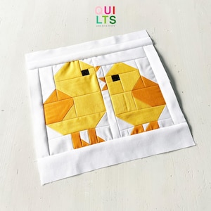 May include: A handmade quilt block featuring two yellow and orange geometric chicks with black eyes. The chicks are set against a white background and framed by a white border. The text "QUILTS" and "AND NICE STUFF" is visible in the upper left corner.