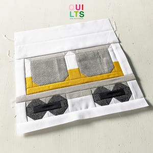 May include: A geometric quilt block featuring a train design. The block uses white, gray, yellow, and black fabric. The top of the block has the text "QUILTS AND NICE STUFF" in green, red, and white.
