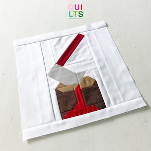 Peut inclure: Un bloc de courtepointe fait à la main avec un motif géométrique. Il présente des formes rouges, marron, grises et beiges sur un fond blanc. Le texte "QUILTS AND NICE STUFF" est visible dans le coin supérieur droit.