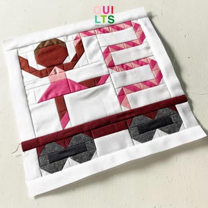 Könnte beinhalten: Ein handgefertigter Quiltblock mit einer Ballerina in einem rosa Tutu und einem geometrischen Design. Der Quiltblock hat einen weißen Hintergrund mit einem rot-rosa bandartigen Design. Der Text "QUILTS AND NICE STUFF" befindet sich oben links.