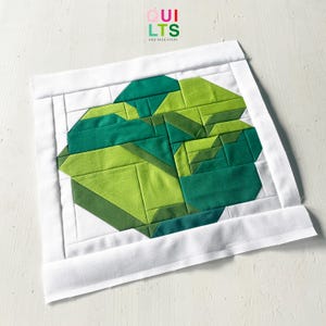 Könnte beinhalten: Ein quadratischer Quiltblock mit einem geometrischen Design in Grüntönen, umgeben von einem weißen Rand. Das Design besteht aus verschiedenen Formen und Winkeln und erzeugt ein modernes, abstraktes Muster. Der Text "QUILTS AND NICE STUFF" befindet sich in der oberen rechten Ecke.