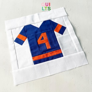 Op de afbeelding: Een wit vierkant van stof met een applicatie van een blauw en oranje voetbalshirt. Het shirt heeft het nummer "4" op de voorkant.