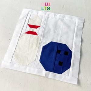 May include: A white fabric square with a blue and white bowling ball and a white bowling pin with red and white stripes. The fabric square has the text "QUI LTS AND NICE STUFF" in the top centre.