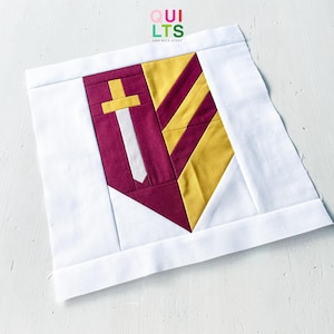 Op de afbeelding: Een vierkant stoffen quiltblok met een centraal schildontwerp dat verticaal is verdeeld. De linkerkant is diepmagenta met een geel en wit zwaardicoon. De rechterkant is voorzien van diagonale gele en magentakleurige strepen.