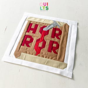 Könnte beinhalten: Ein quadratischer Quiltblock mit dem Wort "ROR" in roten Buchstaben, vor einem braunen und beigen Hintergrund. Der Block ist mit einem weißen Rand versehen. Der Text "QUILTS AND NICE STUFF" befindet sich in der oberen linken Ecke.