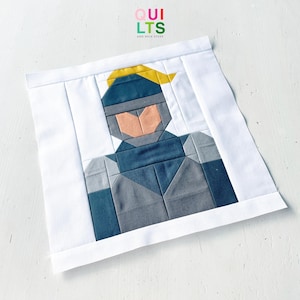 Op de afbeelding: Een vierkant quiltblok van stof met een samengesteld ontwerp van een gestileerd figuur met een geel haarelement, een perzikkleurig gezicht en donkerblauwe en grijze kleding, omlijst door een witte rand. De tekst QUILTS AND NICE STUFF is zichtbaar.