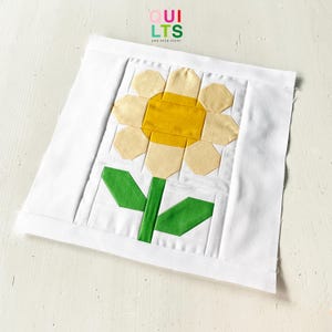 Peut inclure: Un bloc de courtepointe carré avec un motif de fleur géométrique. La fleur a un centre jaune, des pétales jaune clair, une tige et des feuilles vertes, le tout sur un fond blanc. Le texte "QUILTS AND NICE STUFF" est dans le coin supérieur droit.