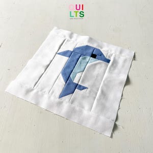 May include: A blue and white fabric dolphin is sewn onto a white fabric square. The fabric square is surrounded by a white border. The text "QUILTS AND NICE STUFF" is printed in rainbow colors at the top of the image.