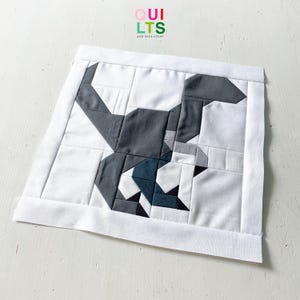 May include: A handmade quilt square featuring a geometric design of a dog. The dog is depicted in shades of gray, white, and a touch of blue. The square is bordered with a white fabric and the text "QUILTS AND NICE STUFF" is in the top right corner.