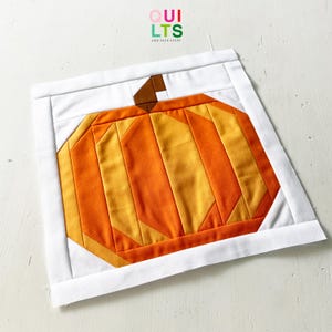 Könnte beinhalten: Ein handgefertigter, gesteppter Kürbisblock mit weißem Rand. Der Kürbis besteht aus orangefarbenen und gelben Stoffstücken mit einem braunen Stiel. Der Text "QUILTS AND NICE STUFF" befindet sich in der oberen rechten Ecke.