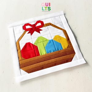 Könnte beinhalten: Ein gestepptes Osternest-Design mit einer roten Schleife und bunten Eiern. Der Korb ist braun, und die Eier sind rot, grün, blau und gelb. Das Design ist mit einem weißen Rand versehen. Der Text "QUILTS AND NICE STUFF" befindet sich oben rechts.