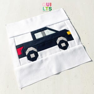May include: A fabric quilt block featuring a geometric design of a dark blue car with gray, red, and yellow accents. The car is set against a white background, with the text "QUILTS AND NICE STUFF" in the upper right corner.