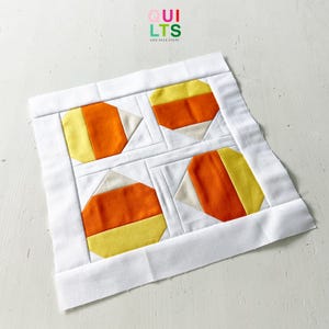 Peut inclure: Un bloc de patchwork fait main avec quatre motifs de bonbons de maïs jaunes, oranges et crème. Les formes de bonbons de maïs sont posées sur un fond blanc et encadrées d'une bordure blanche. Le texte "QUILTS" et "AND NICE STUFF" est dans le coin supérieur droit.