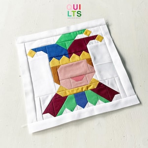 May include: A square fabric quilt block featuring a colorful jester character pieced together from solid cotton segments in blue, green, yellow, maroon, and peach. The jester is centered on a white background with a white border frame.