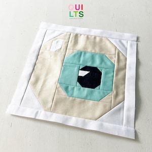Peut inclure: Un bloc de courtepointe carré en tissu avec un motif d'œil géométrique. L'œil est composé de formes bleu clair, noires et blanches, sur un fond beige. Le bloc est encadré d'une bordure blanche. Le texte "QUILTS AND NICE STUFF" est dans le coin supérieur gauche.