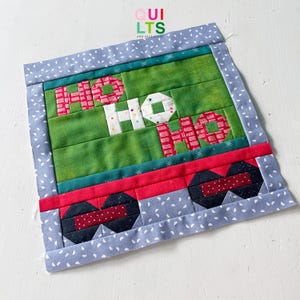 May include: A green and red patchwork quilt block with the word "HO HO HO" in white letters. The block is surrounded by a blue border with white polka dots.
