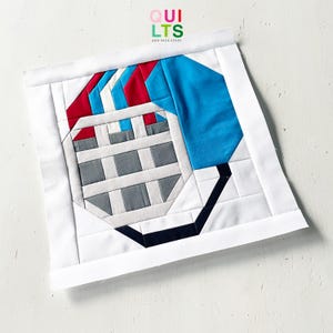 May include: A handmade quilt block featuring a football helmet design. The helmet is composed of blue, red, white, and gray fabric pieces, with a black facemask. The block is bordered by white fabric and includes the text "QUILTS AND NICE STUFF".