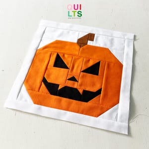 Könnte beinhalten: Ein handgefertigter Halloween-Quiltblock mit einem orangefarbenen Kürbis mit einem geschnitzten Gesicht. Der Kürbis ist vor einem weißen Hintergrund platziert und mit einem weißen Rand umrahmt. Der Text "QUILTS AND NICE STUFF" steht oben.