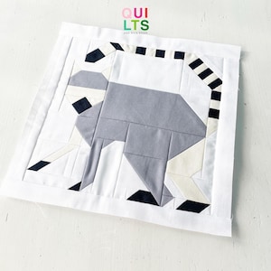 May include: A handmade quilt square featuring a geometric lemur design. The lemur is depicted in shades of gray, black, and white, with a striped tail. The quilt square is bordered by white fabric. The text "QUILTS AND NICE STUFF" is in the top right corner.