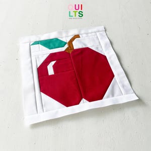 Könnte beinhalten: Ein gestepptes Apfeldesign mit einem roten Apfel, einem grünen Blatt und einem braunen Stiel. Der Apfel ist vor einem weißen Hintergrund mit einem weißen Rand platziert. Der Text "QUILTS AND NICE STUFF" befindet sich in der oberen rechten Ecke.