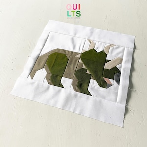 Könnte beinhalten: Ein grüner und brauner Patchwork-Quiltblock, der einen Triceratops-Dinosaurier darstellt. Der Block besteht aus verschiedenen Stoffquadraten, die zusammengenäht wurden, um die Form des Dinosauriers zu bilden. Der Quiltblock befindet sich auf einem weißen Hintergrund.