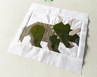 Pode incluir: Um bloco de colcha de retalhos verde e castanho que representa um dinossauro triceratops. O bloco é feito de vários quadrados de tecido costurados juntos para criar a forma do dinossauro. O bloco de colcha está sobre um fundo branco.