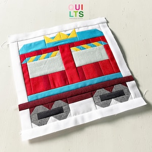 May include: A colorful, handmade quilt block featuring a stylized fire truck design. The quilt block is primarily red, with blue, yellow, gray, and white accents. The text "QUILTS AND NICE STUFF" is in the upper right corner.