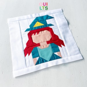 Pode incluir: Um bloco de colcha quadrado de tecido com um design de peças de um personagem com cabelo vermelho, um chapéu de bruxa azul-petróleo com um detalhe amarelo e um corpete azul-petróleo sobre um fundo branco com bordas brancas.