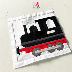 Könnte beinhalten: Ein handgefertigtes Quiltquadrat mit einem Zugdesign. Der Zug ist hauptsächlich schwarz mit roten und grauen Akzenten, vor einem weißen Hintergrund. Der Text "QUILTS AND NICE STUFF" ist oben links sichtbar.