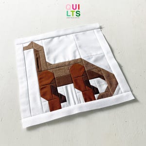 May include: A handmade quilt block featuring a brown and white dinosaur design. The dinosaur is pieced together with various shades of brown fabric, set against a white background. The block is framed with a white border, and the text "QUILTS AND NICE STUFF" is visible.