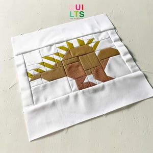 Könnte beinhalten: Ein weißer Stoffquadrat mit einem braunen und gelben Patchwork-Dinosaurier-Design in der Mitte. Der Dinosaurier hat einen gestreiften Rücken und einen braunen Körper. Der Stoff ist gesteppt und hat einen weißen Hintergrund.
