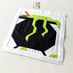 Könnte beinhalten: Ein quadratischer Quiltblock mit einem schwarzen Kessel mit limettengrünen Akzenten und einem weißen Rand. Der Quiltblock hat den Text "QUILTS AND NICE STUFF" in der oberen rechten Ecke.