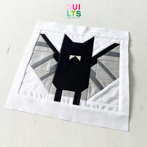 Puede incluir: Un bloque de edredón hecho a mano con un diseño de murciélago negro con pajarita blanca, sobre un fondo geométrico gris y blanco. El bloque está enmarcado con un borde blanco. El texto "QUILTS AND NICE STUFF" está en la esquina superior derecha.